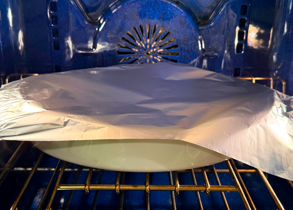 White baking dish loosely covered with aluminum foil and sitting in a blue oven.