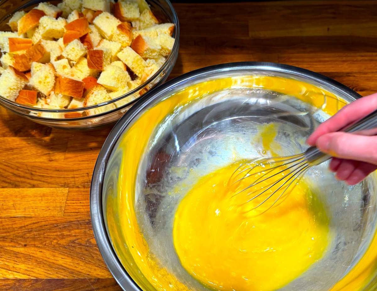 Eggs being beaten with a whisk in a large steel bowl next to a medium glass bowl full of bread cubes.