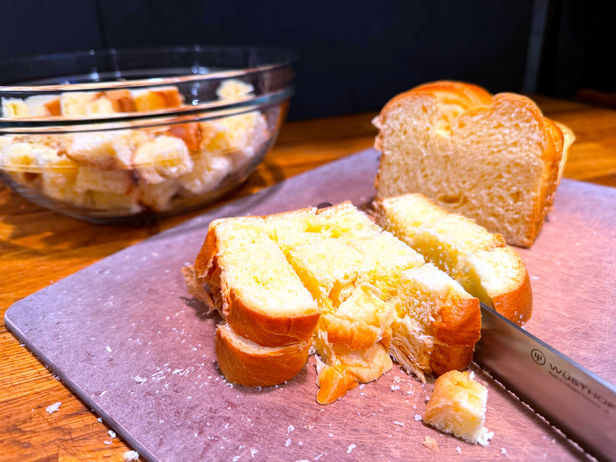 Slices of bread being cut into cubes with a knife on a brown cutting board in front of a glass bowl of bread cubes.