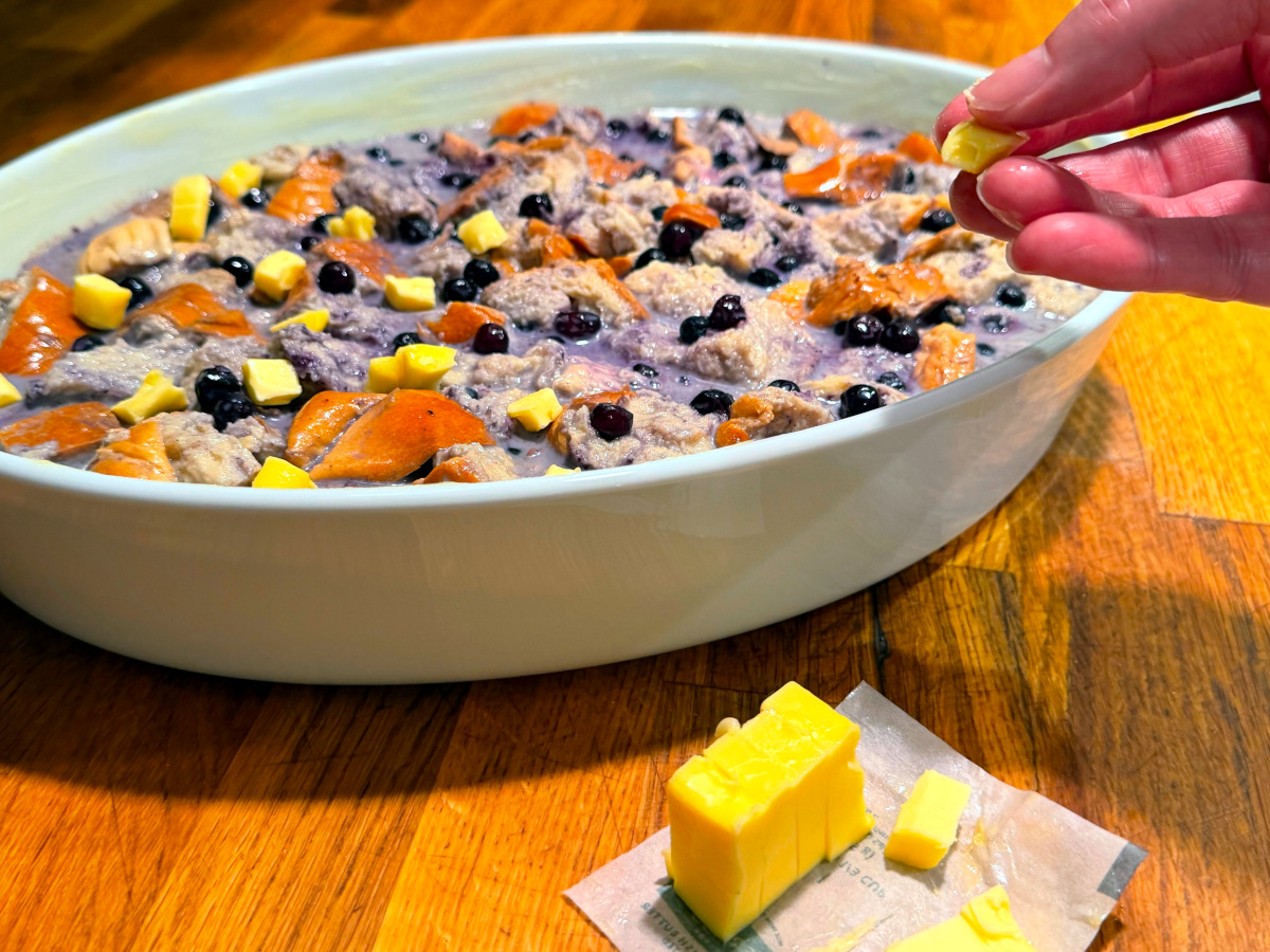 Small bits of butter being placed on top of wet bread cube and blueberry mixture in a white oval baking dish.