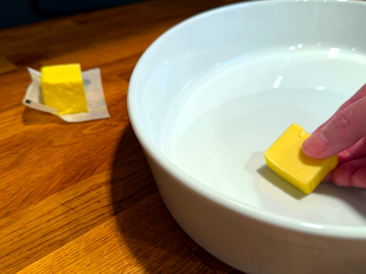 Pat of butter being placed into a white baking dish next to a small square of butter sitting on its wrapper.