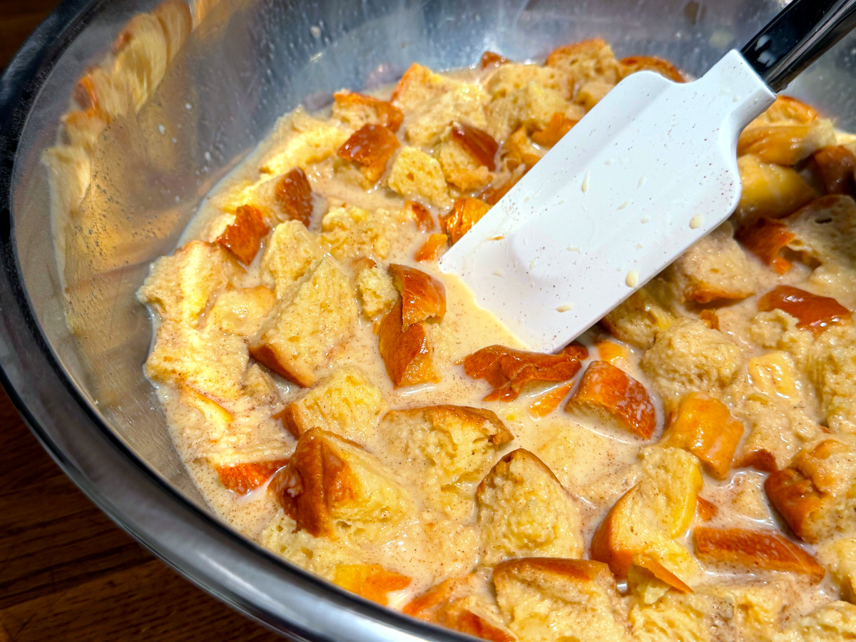 Bread cubes being stirred into yellow liquid with a white silicone spatula in a steel bowl.