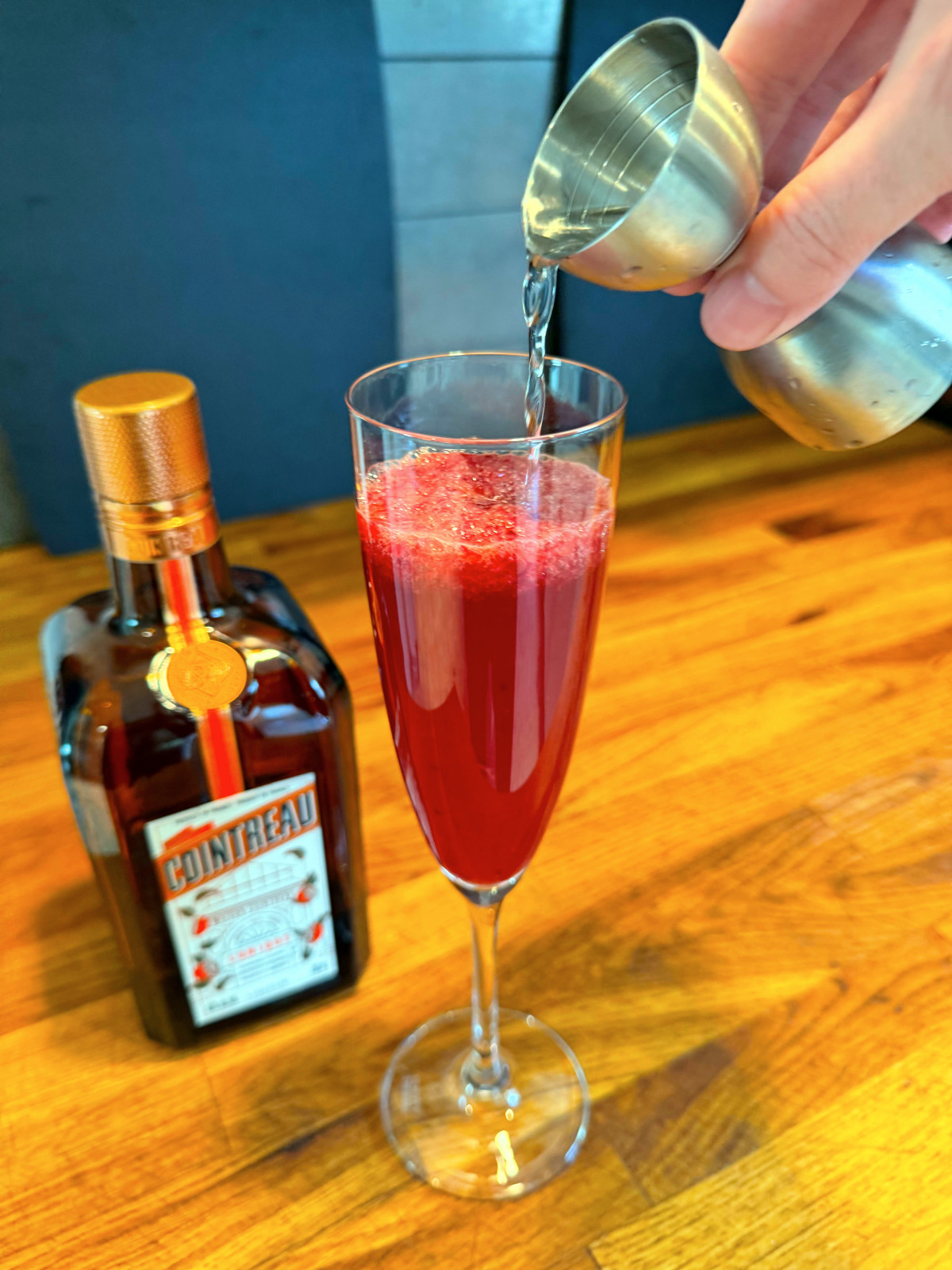 Tipping clear liquid from steel jigger into champagne flute containing red liquid next to a bottle of Cointreau.