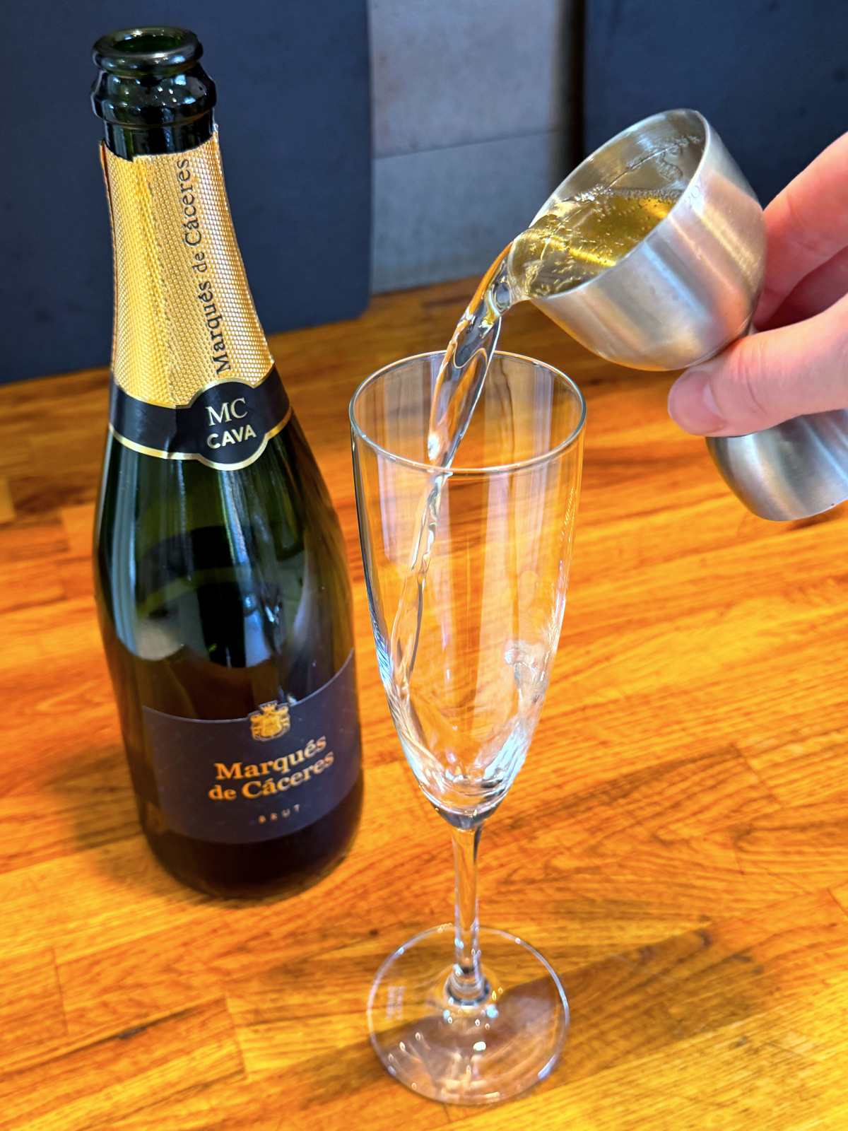 Tipping clear liquid from steel jigger into champagne flute next to a bottle of cava.
