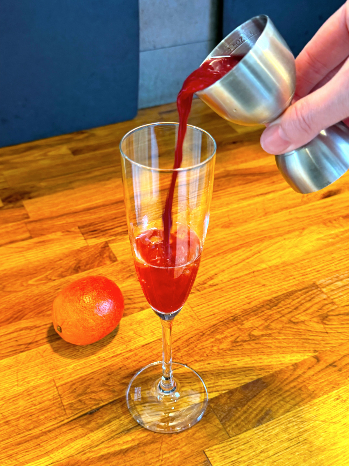 Tipping red liquid from a steel jigger into a champagne flute next to a blood orange.