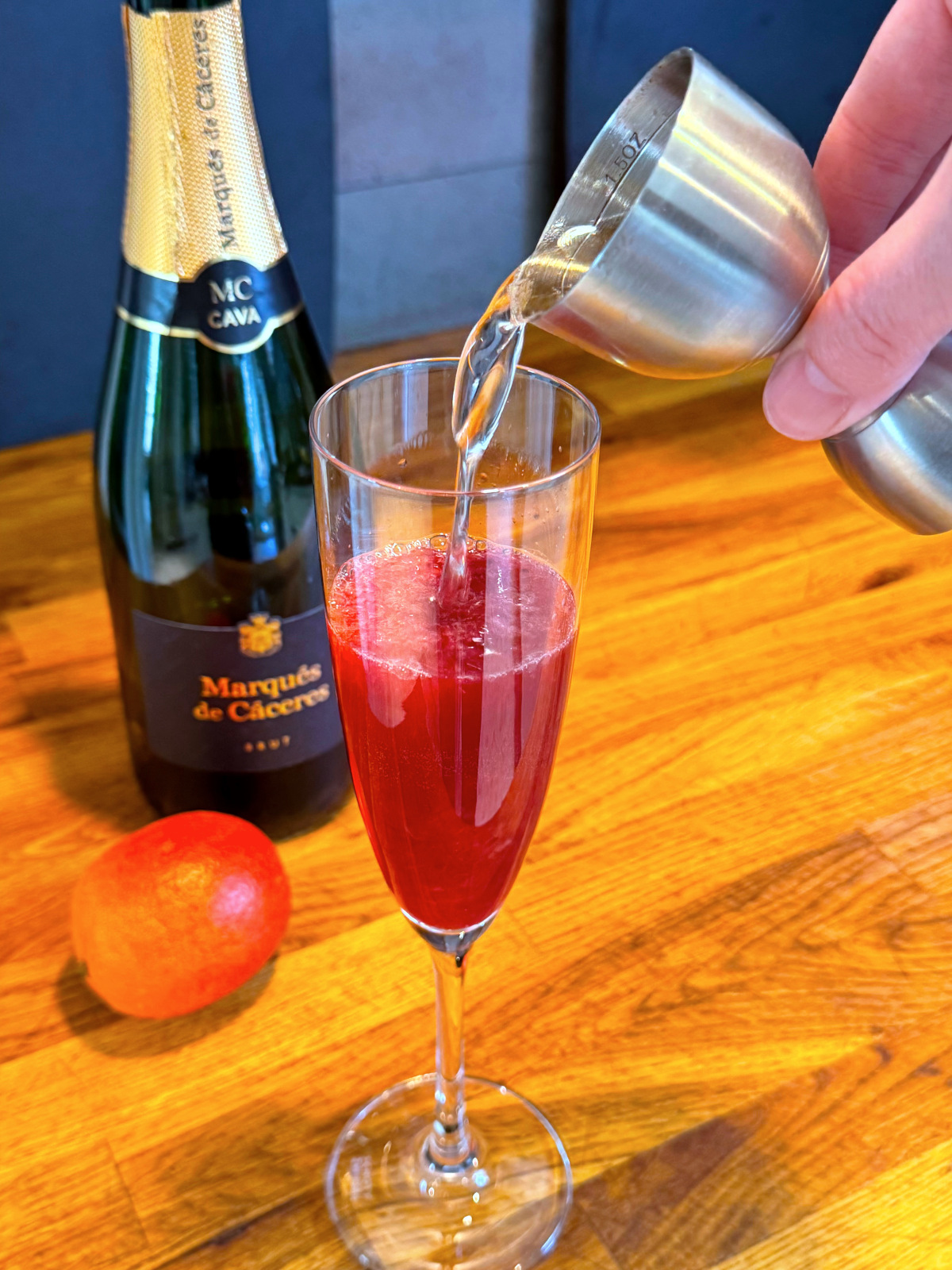 Tipping clear liquid from steel jigger into champagne flute containing red liquid next to a bottle of cava and a blood orange.