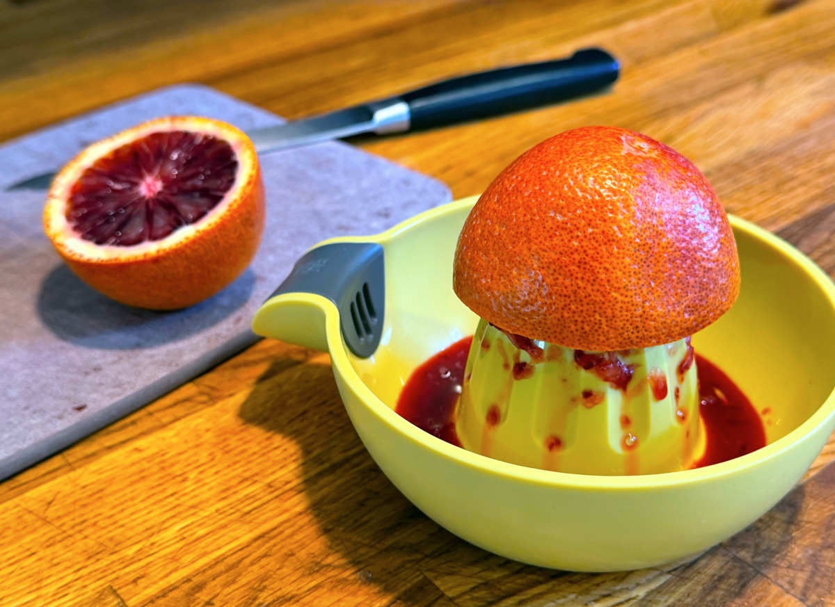Orange half perched on the center of a yellow plastic juicer next to a blood orange half and a knife sitting on a small cutting board.