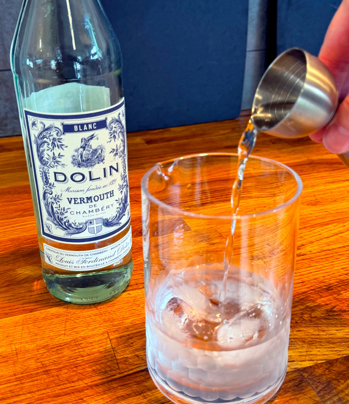 Clear liquid being tipped from a steel measuring jigger into a mixing glass with ice next to a bottle of blanc vermouth.