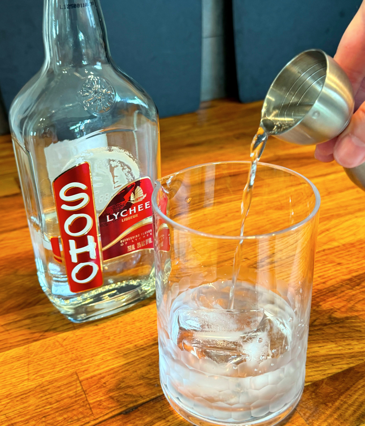 Clear liquid being tipped from a steel jigger into a mixing glass with ice next to a bottle of lychee liqueur.