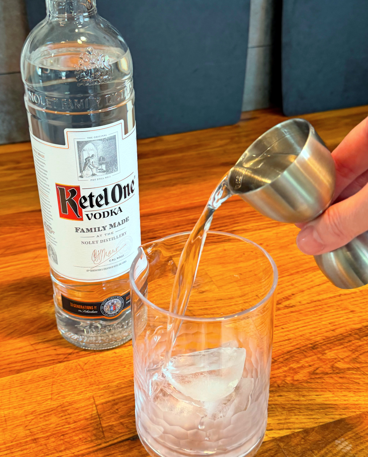 Clear liquid being poured from a steel measuring jigger into a mixing glass with ice next to a bottle of vodka.