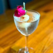 Lychee martini served in a Nick and Nora glass, garnished with a lychee and a dark pink rosebud.
