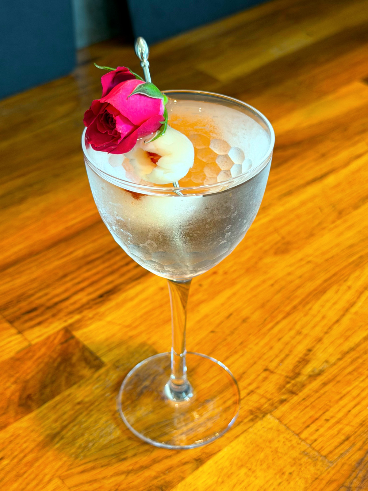Lychee martini served in a Nick and Nora glass, garnished with a lychee and a dark pink rosebud.