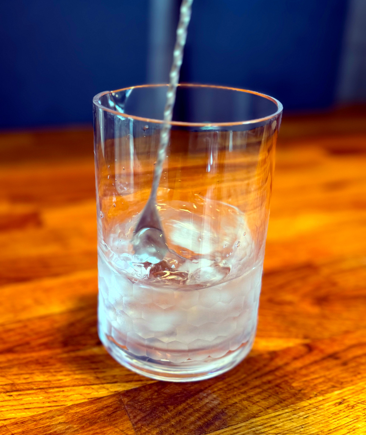 Clear liquid and ice being stirred in a mixing glass with a long metal spoon.