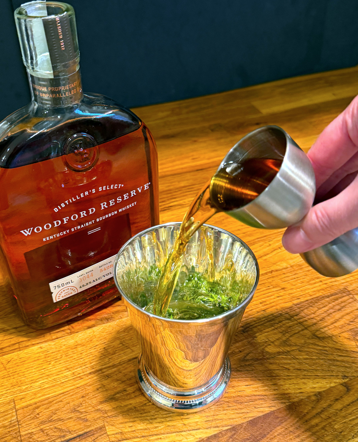 Brown liquid being poured from a steel jigger into a silver julep cup next to a bottle of bourbon.