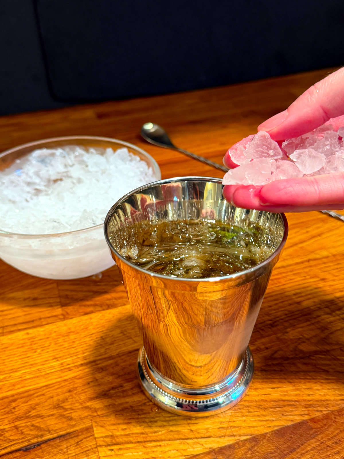 Handful of crushed ice being held over a silver julep cup next to a glass bowl of crushed ice and a bar spoon.