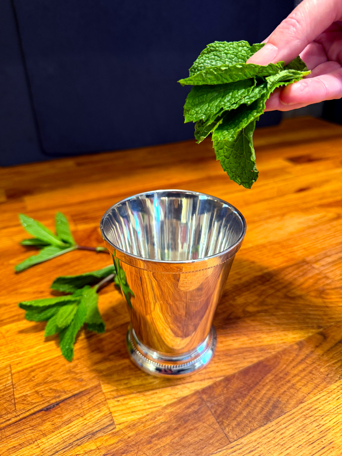 Mint leaves being held over a silver julep cup next to a couple sprigs of mint.