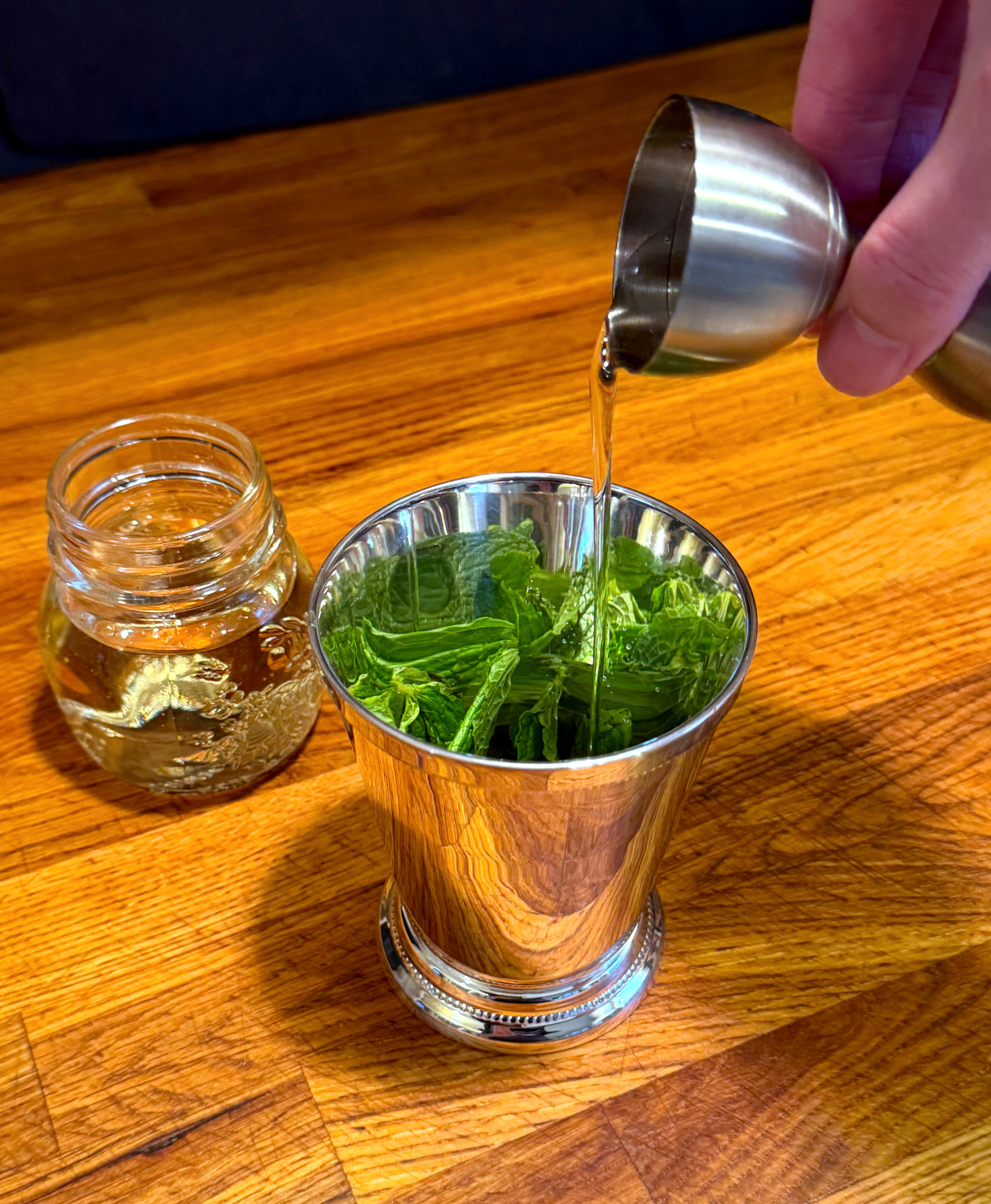 Clear liquid being poured from a steel jigger into a silver julep cup with mint leaves in the bottom next to a small jar of clear liquid.