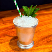 Mint julep served in a silver julep cup garnished with a sprig of mint next to a green and white striped straw.