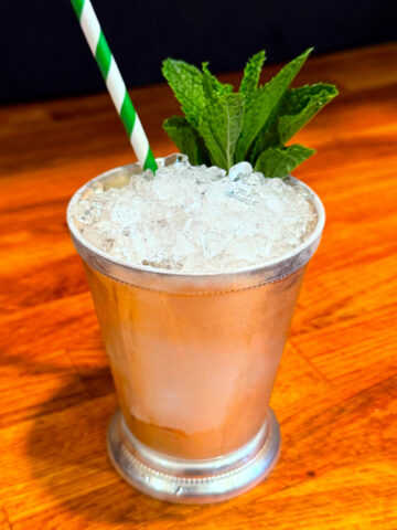 Mint julep served in a silver julep cup garnished with a sprig of mint next to a green and white striped straw.