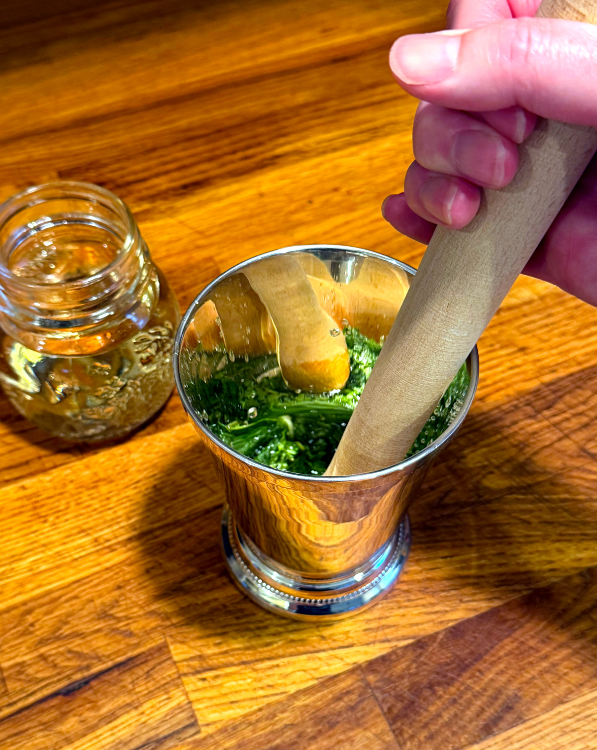 Mint leaves being pressed with a wooden muddler in a silver julep cup next to a small jar of clear liquid.