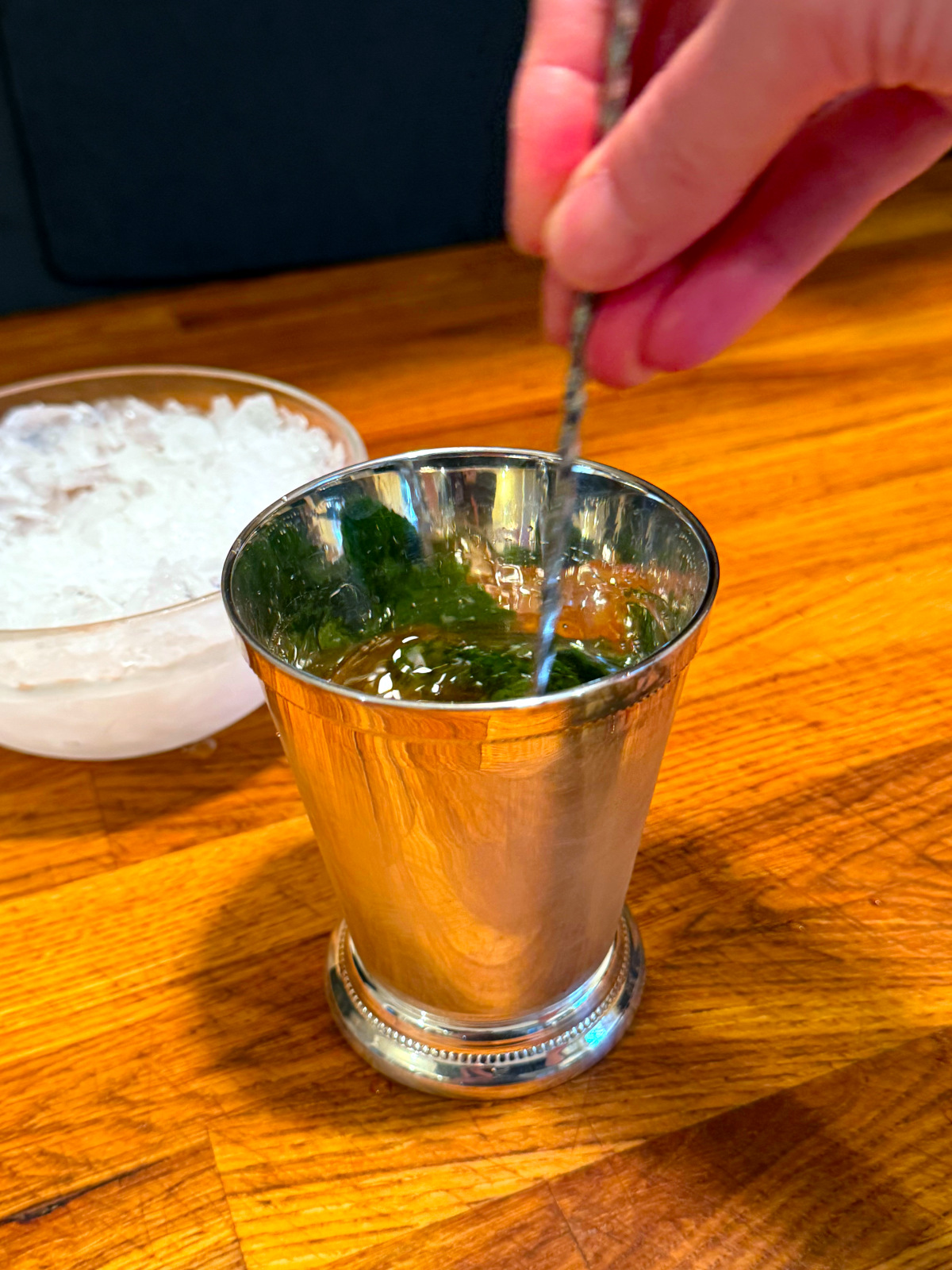 Brown liquid and crushed ice being stirred with a bar spoon in a silver julep cup next to a glass bowl of crushed ice.