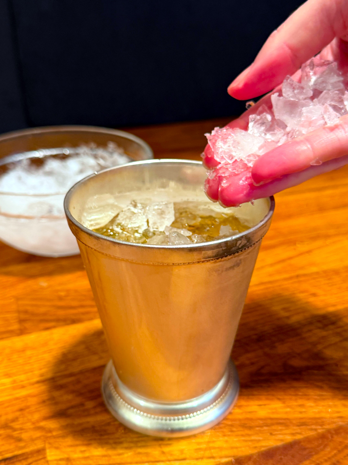 Crushed ice being dropped into a silver julep cup in front of a glass bowl of crushed ice.