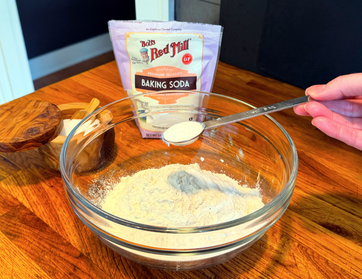 Tipping white powder from a steel measuring spoon into flour in a glass bowl next to a bag of baking soda.