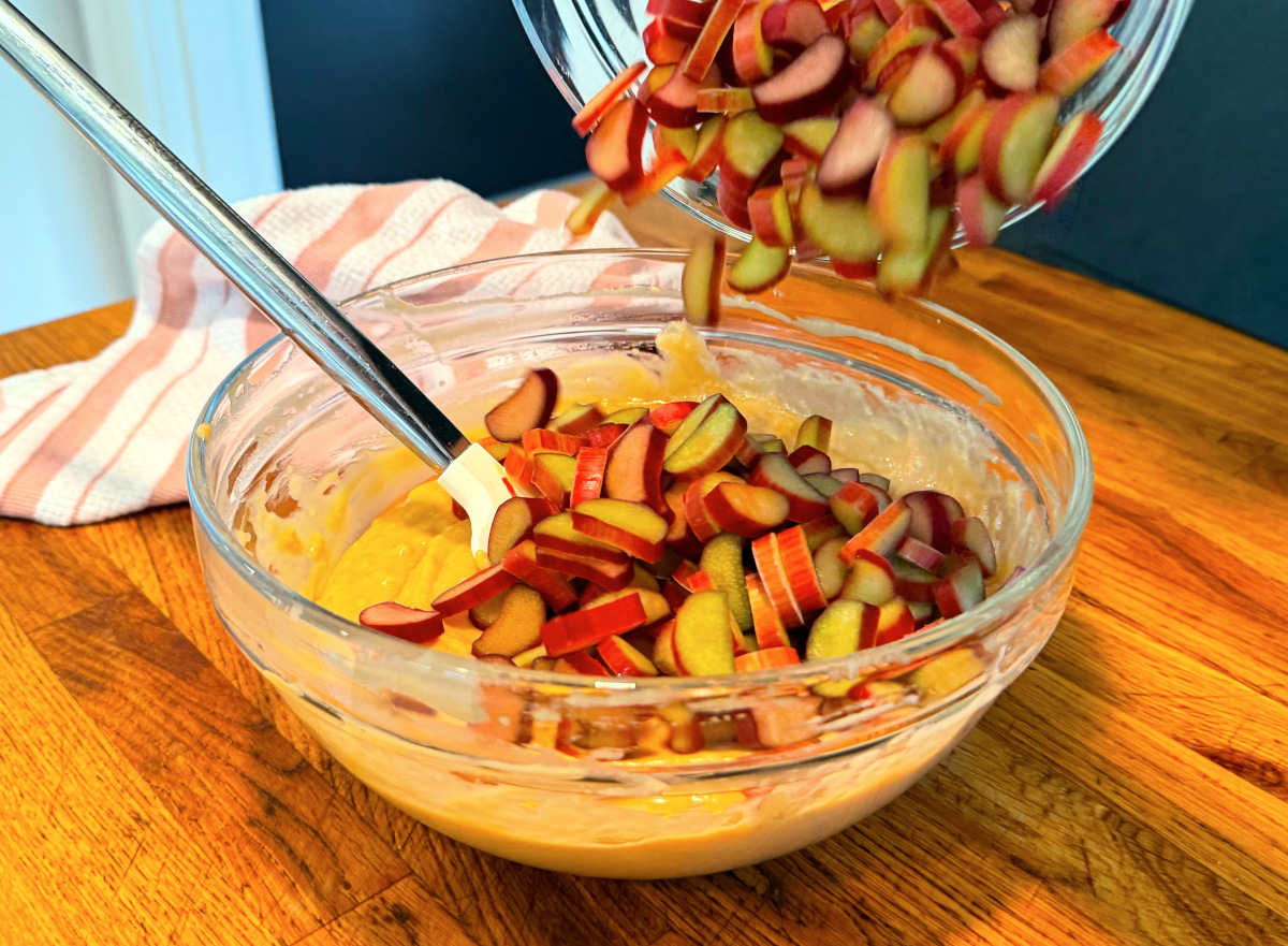 Chopped rhubarb being tipped from a glass bowl into pale brown batter in a larger glass bowl.
