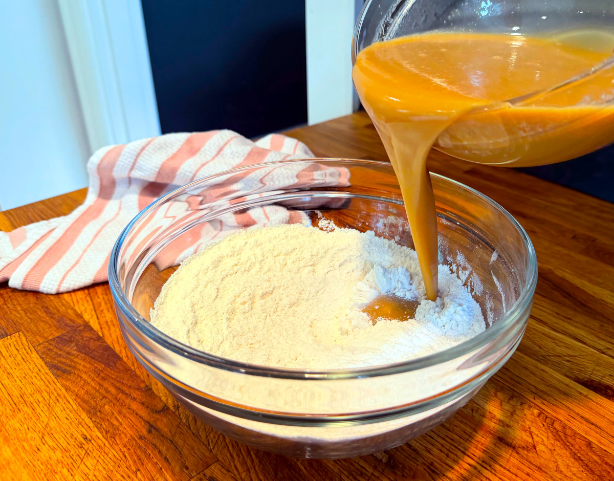 Caramel colored liquid being poured from a glass bowl into dry ingredients in a glass bowl next to a pink and white striped kitchen towel.