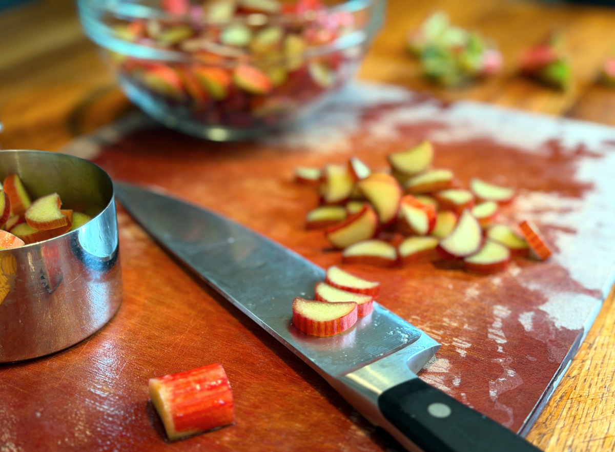 Chopped rhubarb and a chef's knife on a cutting board along with a steel measuring cup and glass bowl containing chopped rhubarb.