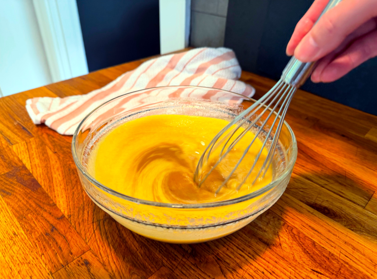 Brown sugar being whisked into yellow liquid in a glass bowl next to a pink and white striped kitchen towel.