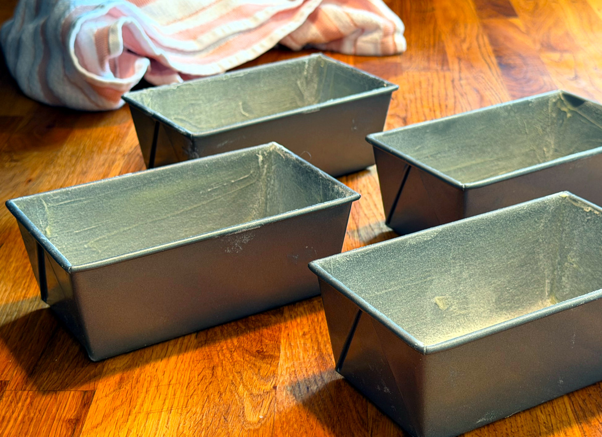 Four small buttered and floured loaf pans sitting on a wood counter.