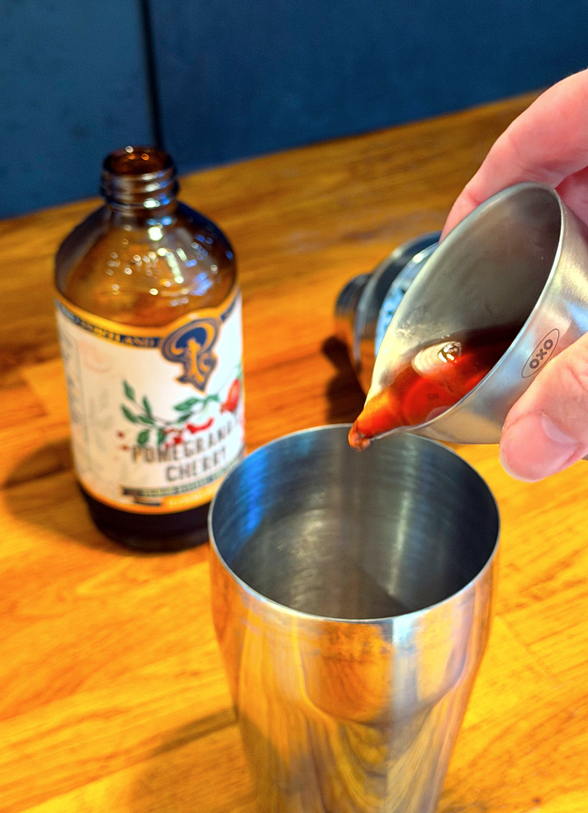 Red liquid being tipped from a steel measuring jigger into a cocktail shaker next to a bottle of grenadine.