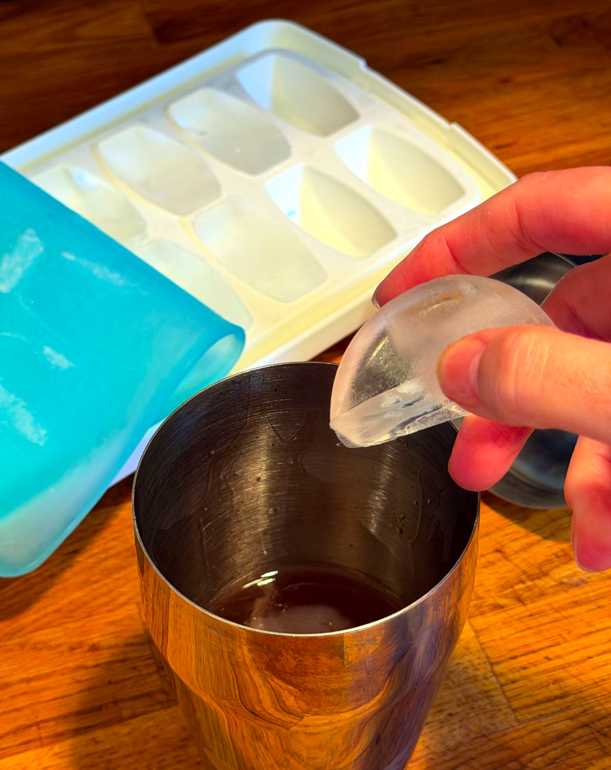 Ice cube being dropped into a steel cocktail shaker next to an ice cube tray.