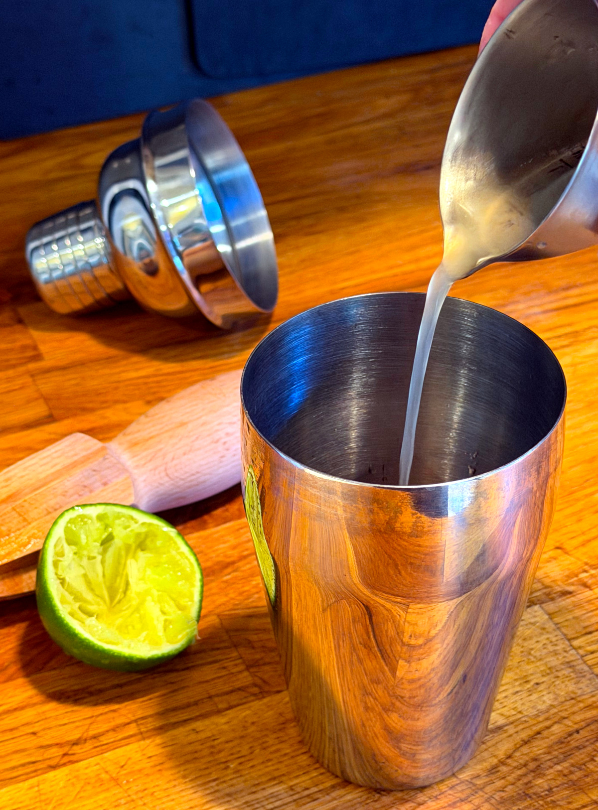 Pale green liquid being poured from a steel measuring jigger into a cocktail shaker next to a juiced lime half and citrus reamer.