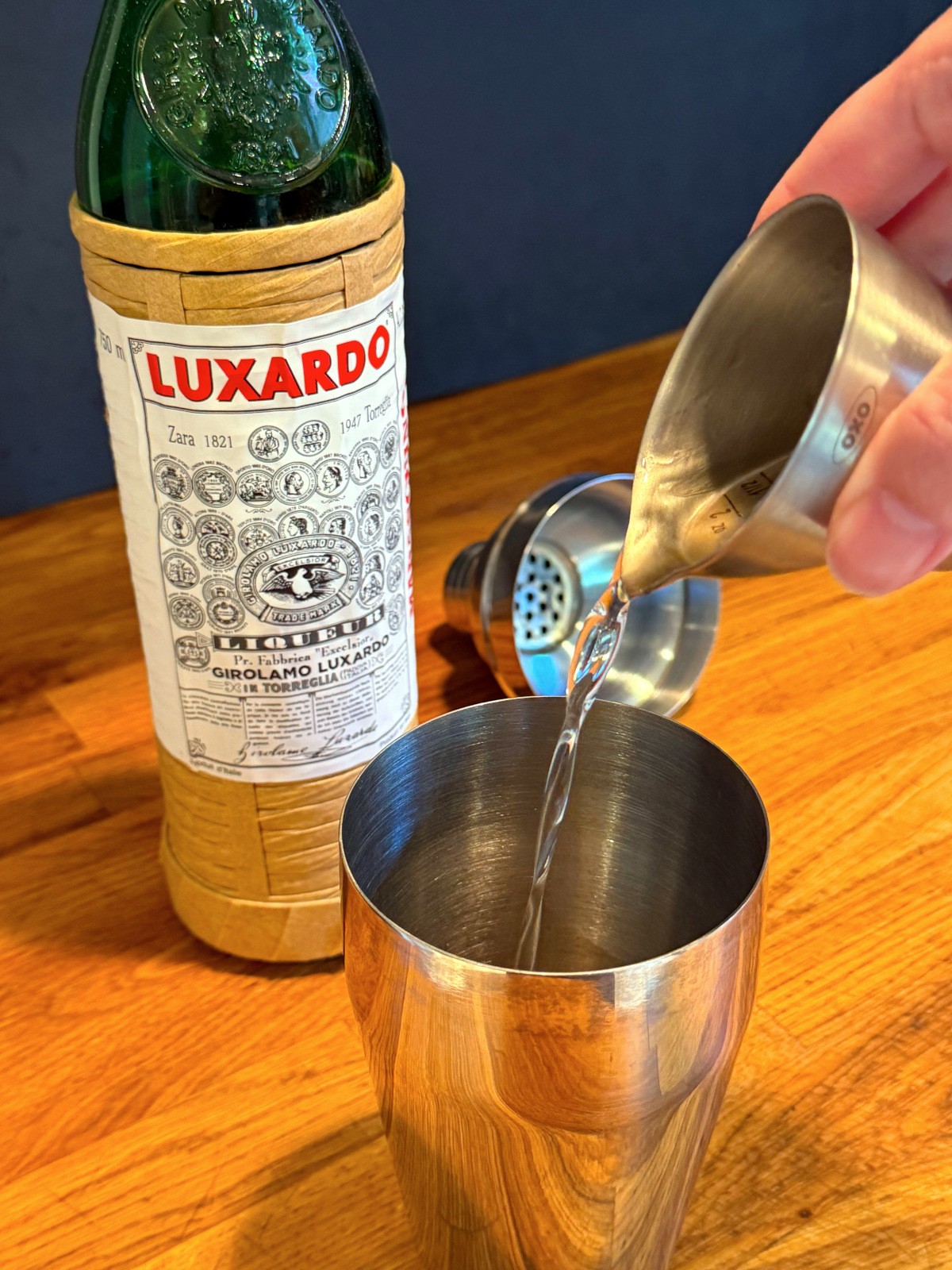 Clear liquid being poured from a steel measuring jigger into a cocktail shaker next to a bottle of maraschino liqueur.