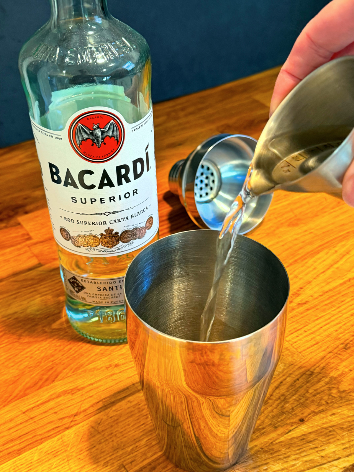 Clear liquid being poured from a steel measuring jigger into a cocktail shaker net to a bottle of white rum.
