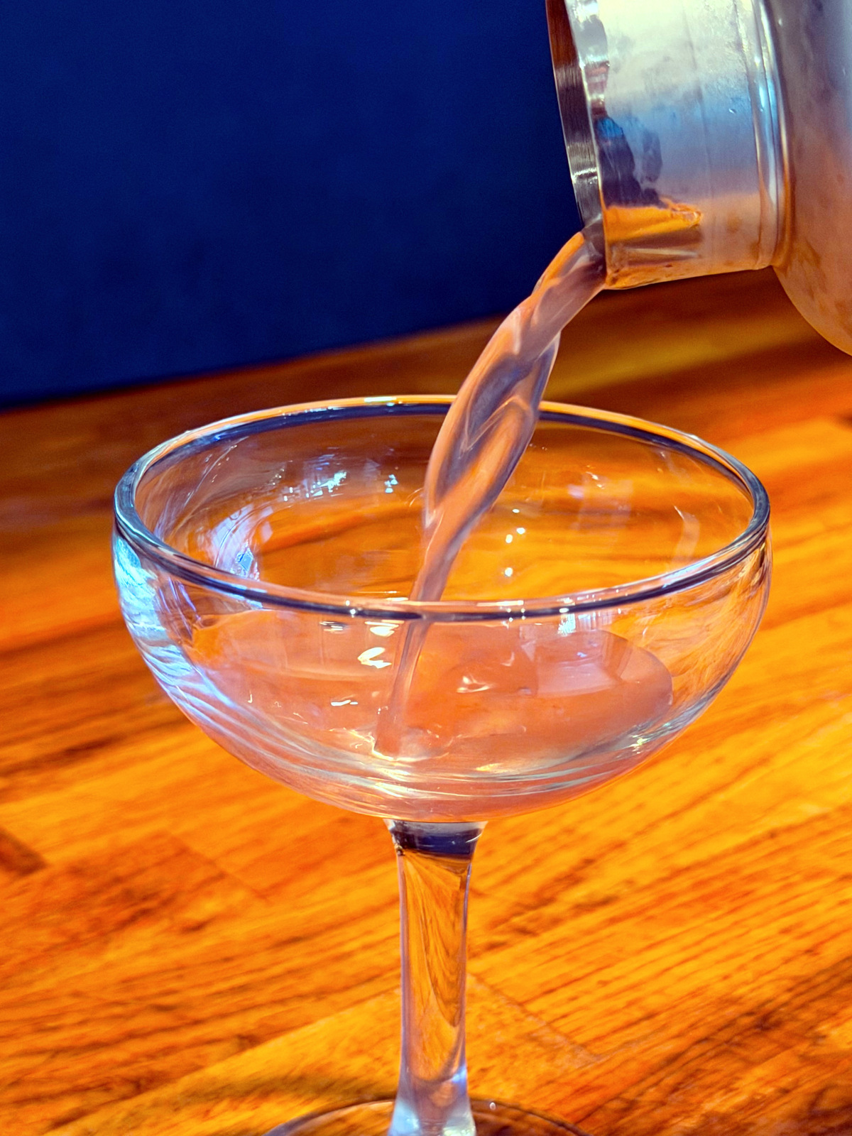 Pale pink liquid being poured from a cocktail shaker into a coupe glass.