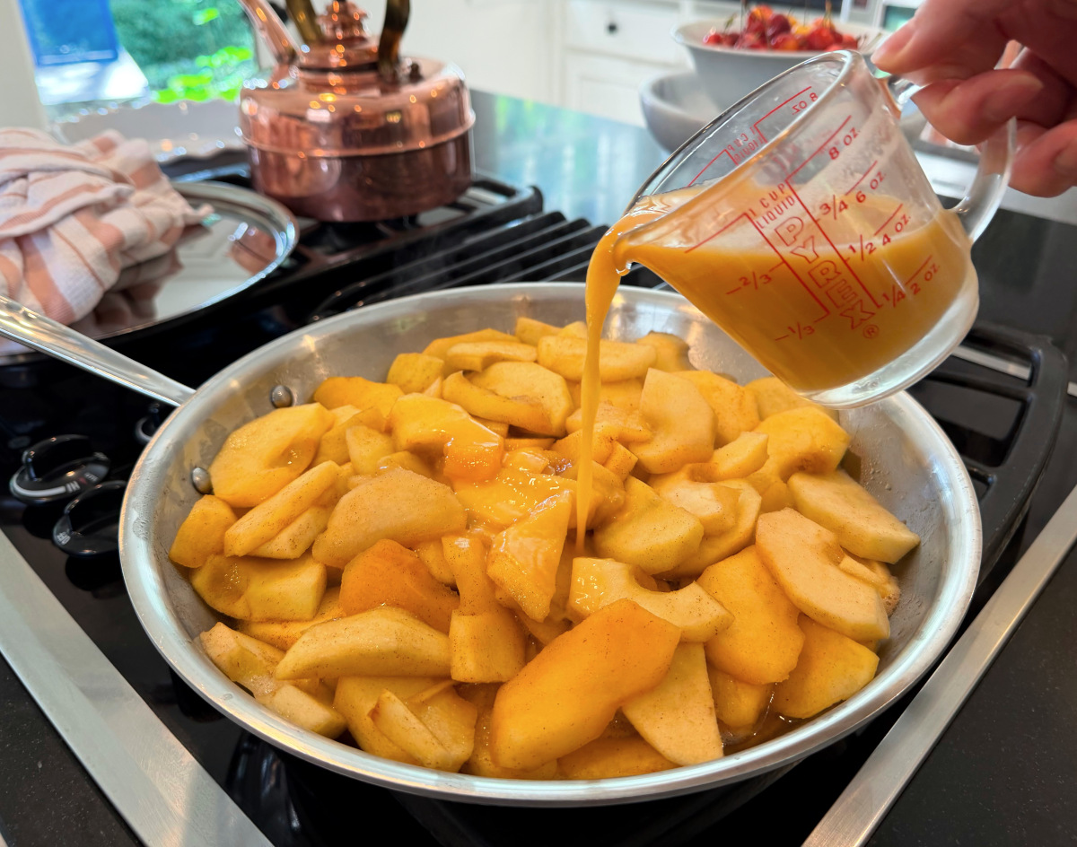 Cloudy amber liquid being poured from a glass measuring cup into a steel skillet full of apple slices.