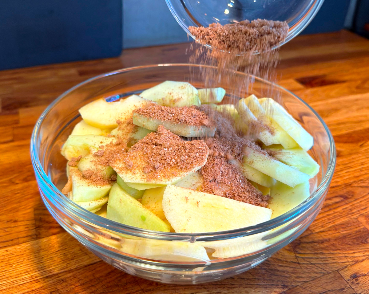 Sugar and cinnamon being tipped from a small glass bowl over sliced apples in a large glass bowl.