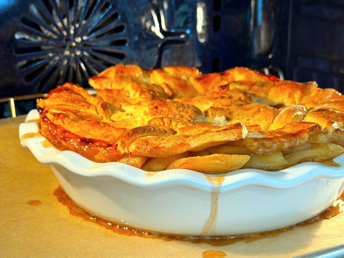Apple pandowdy in a deep white pie dish on a parchment covered baking sheet in a blue oven.