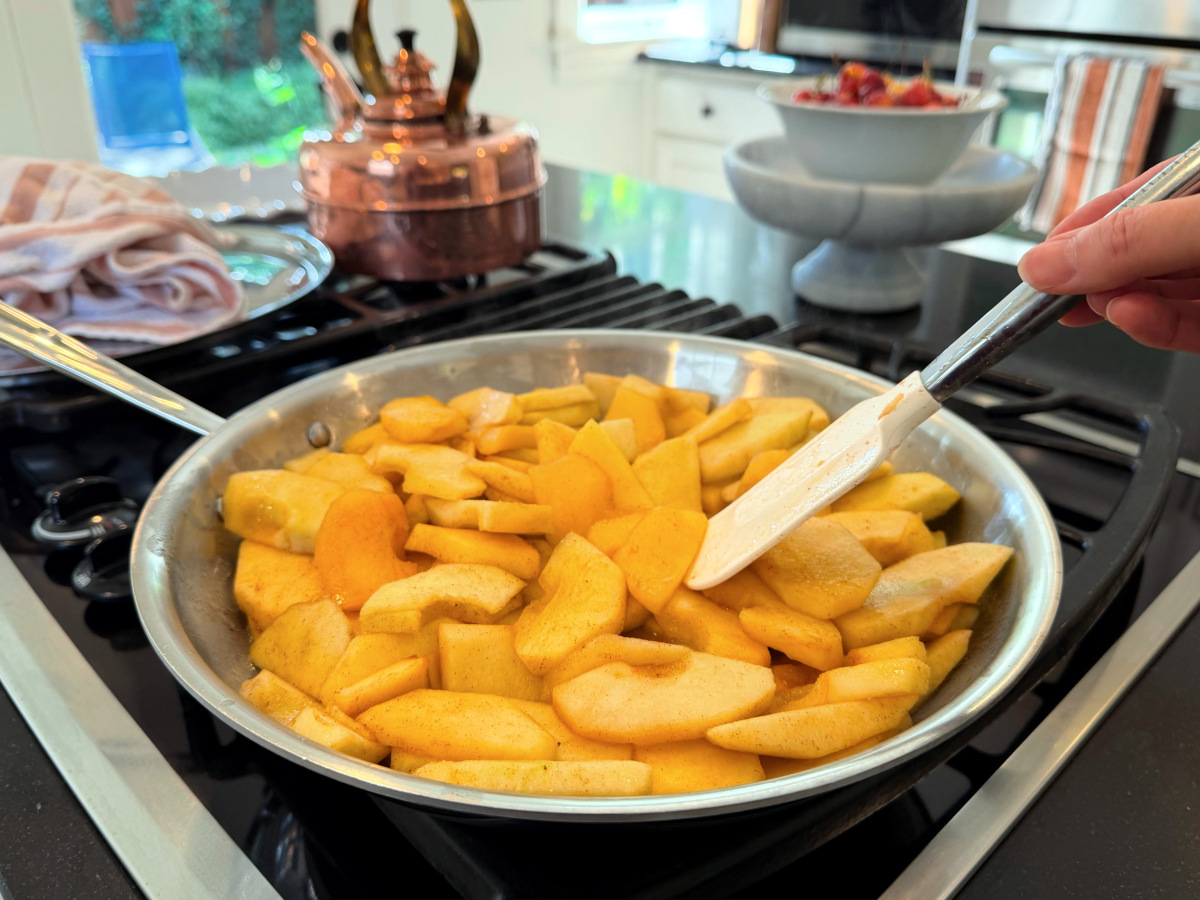 Apple slices in a large steel skillet being stirred with a white silicone spatula over the stove.