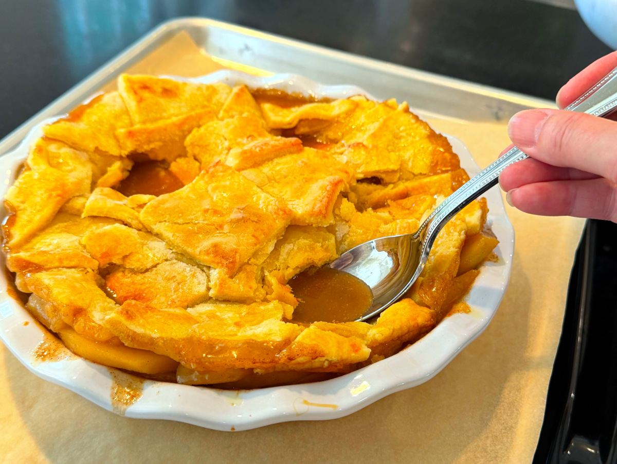 Large silver spoon being pressed down into amber liquid underneath golden pastry squares in a deep white pie dish.