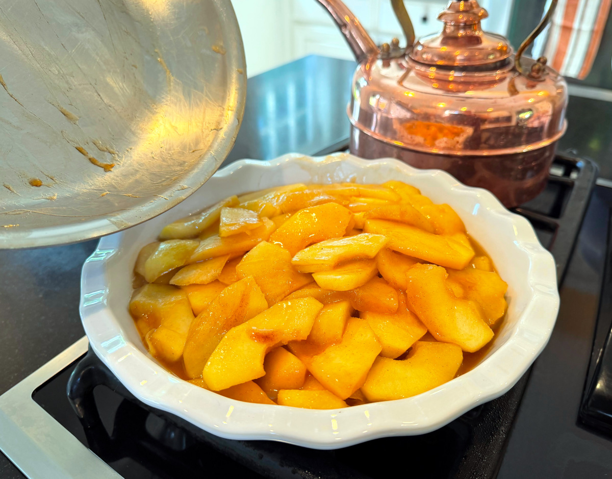 Apples being transferred from a steel skillet into a deep white pie dish sitting next to a copper tea kettle.