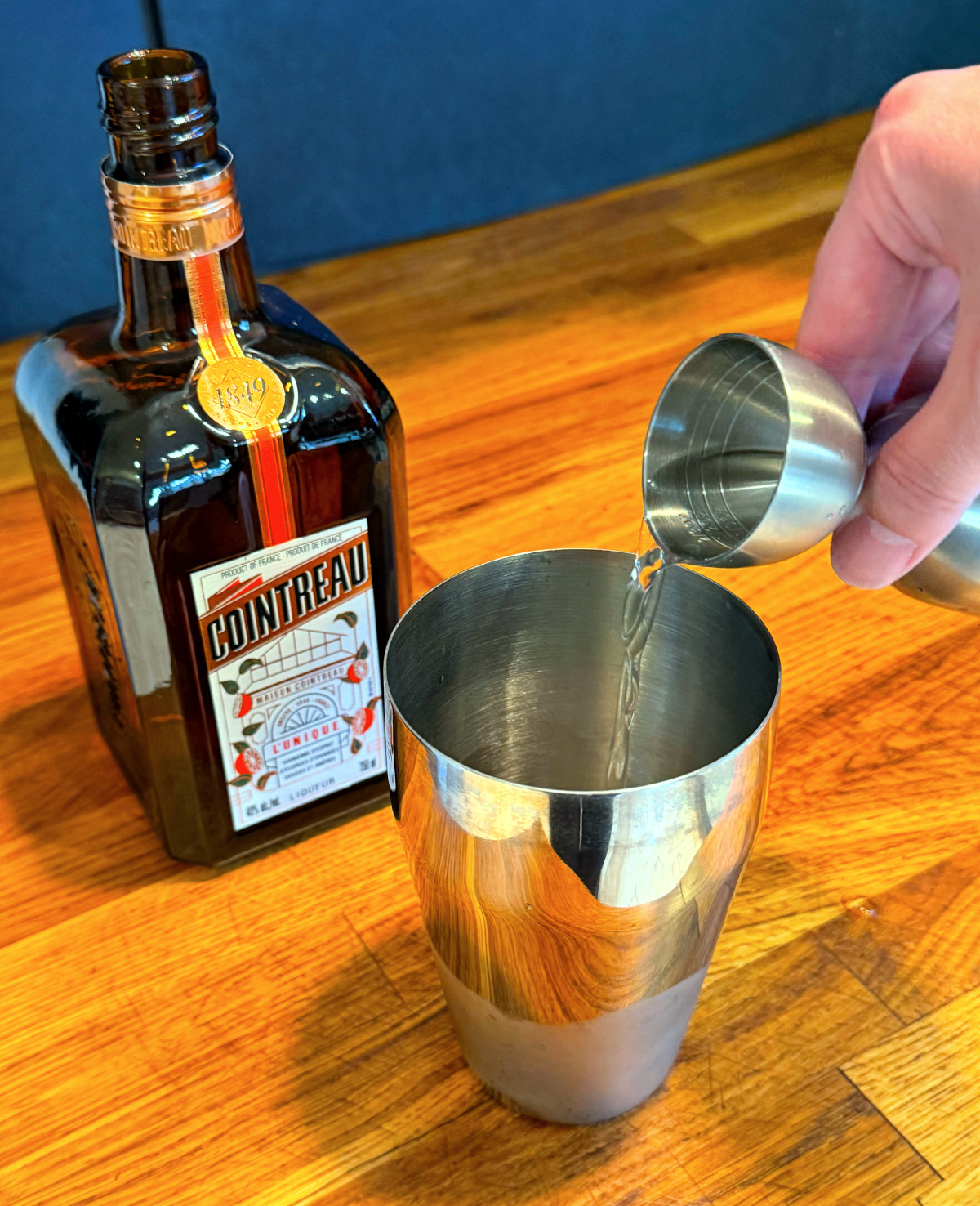Clear liquid being tipped from a steel measuring jigger into a cocktail shaker next to a bottle of Cointreau.
