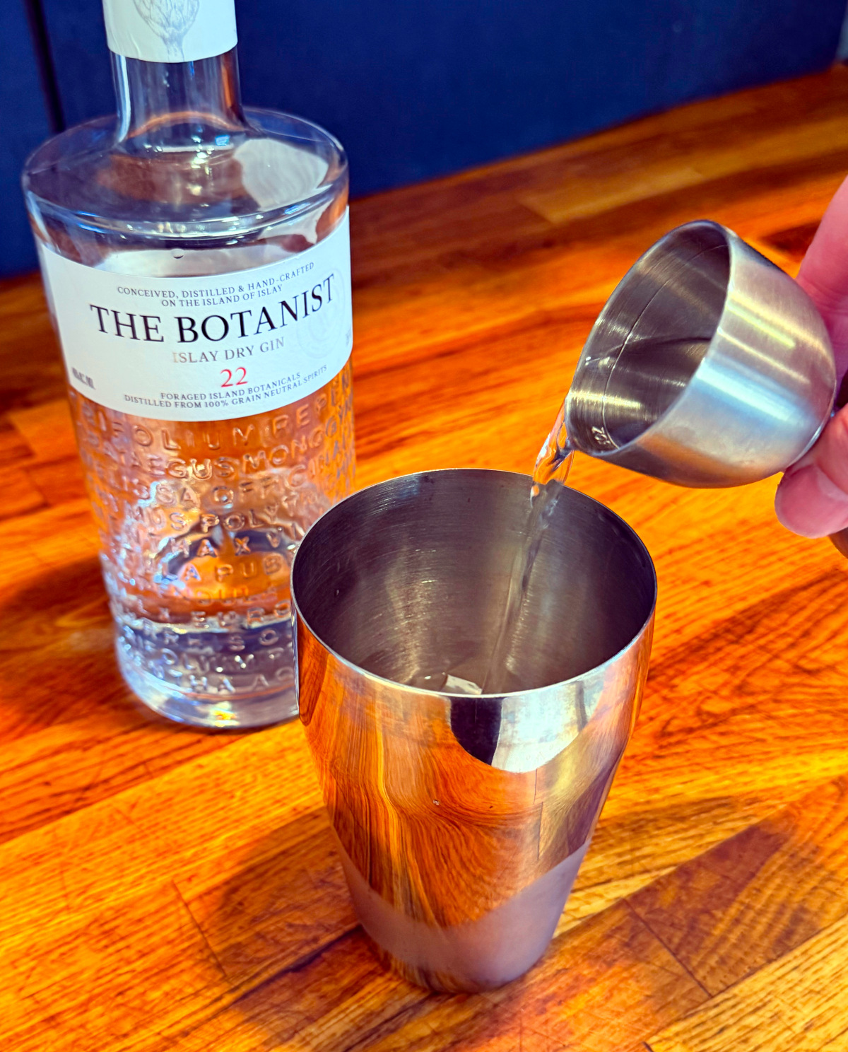 Clear liquid being tipped from a steel measuring jigger into a cocktail shaker next to a bottle of gin.