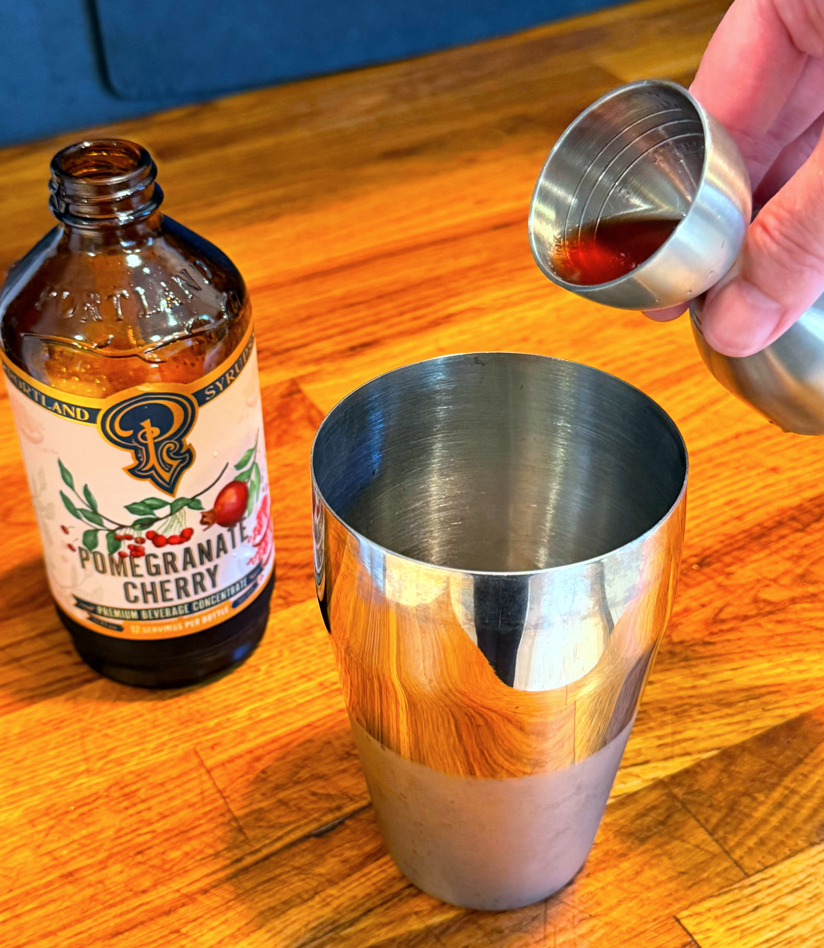 Red liquid being tipped from a steel measuring jigger into a cocktail shaker next to a bottle of grenadine.