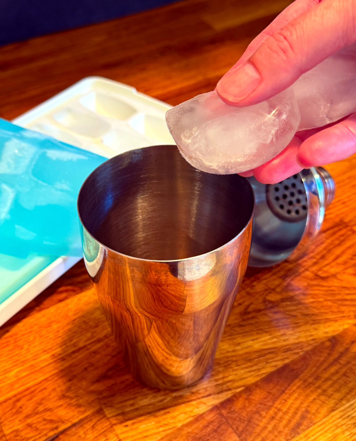 Ice cubes being dropped into a cocktail shaker next to a blue and white ice cube tray.