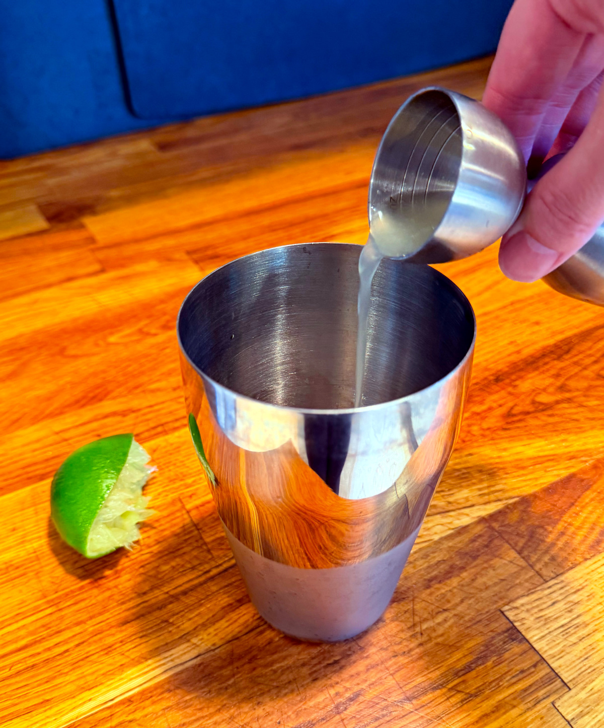 Pale green liquid being tipped from a steel measuring jigger into a cocktail shaker next to a spent lime half.