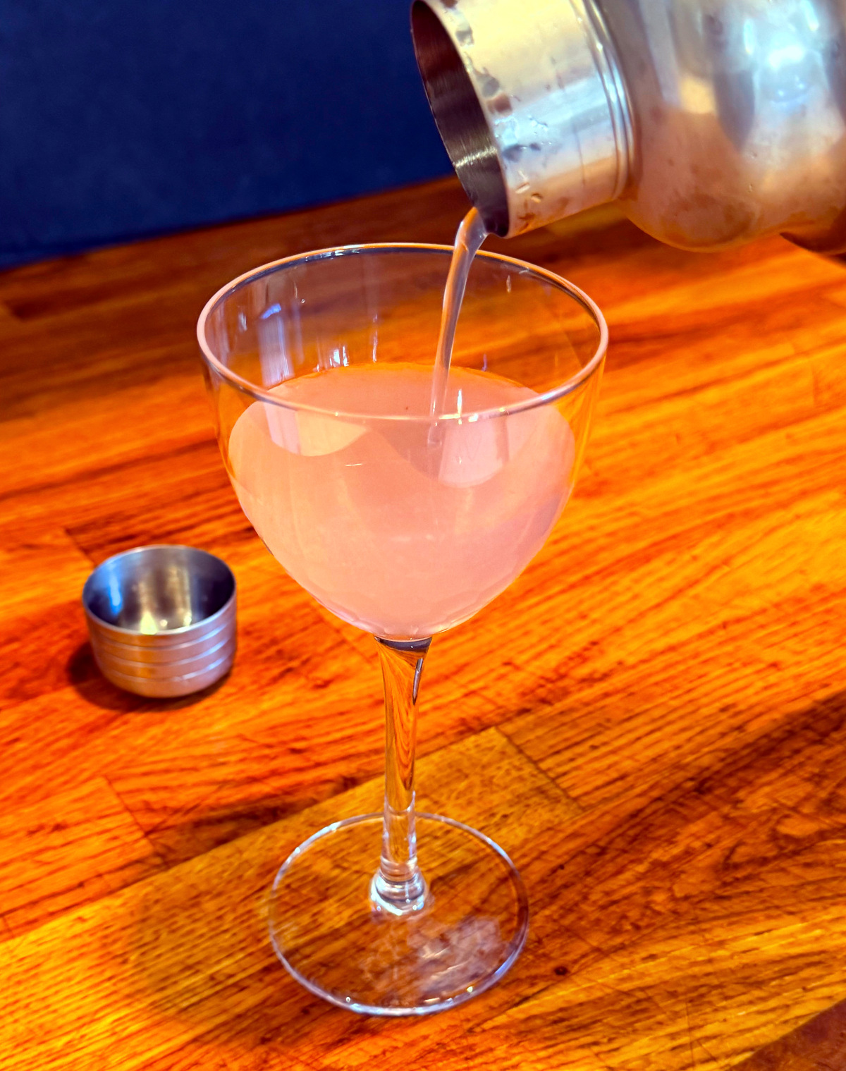 Pale pink liquid being poured from a cocktail shaker into a Nick and Nora glass.